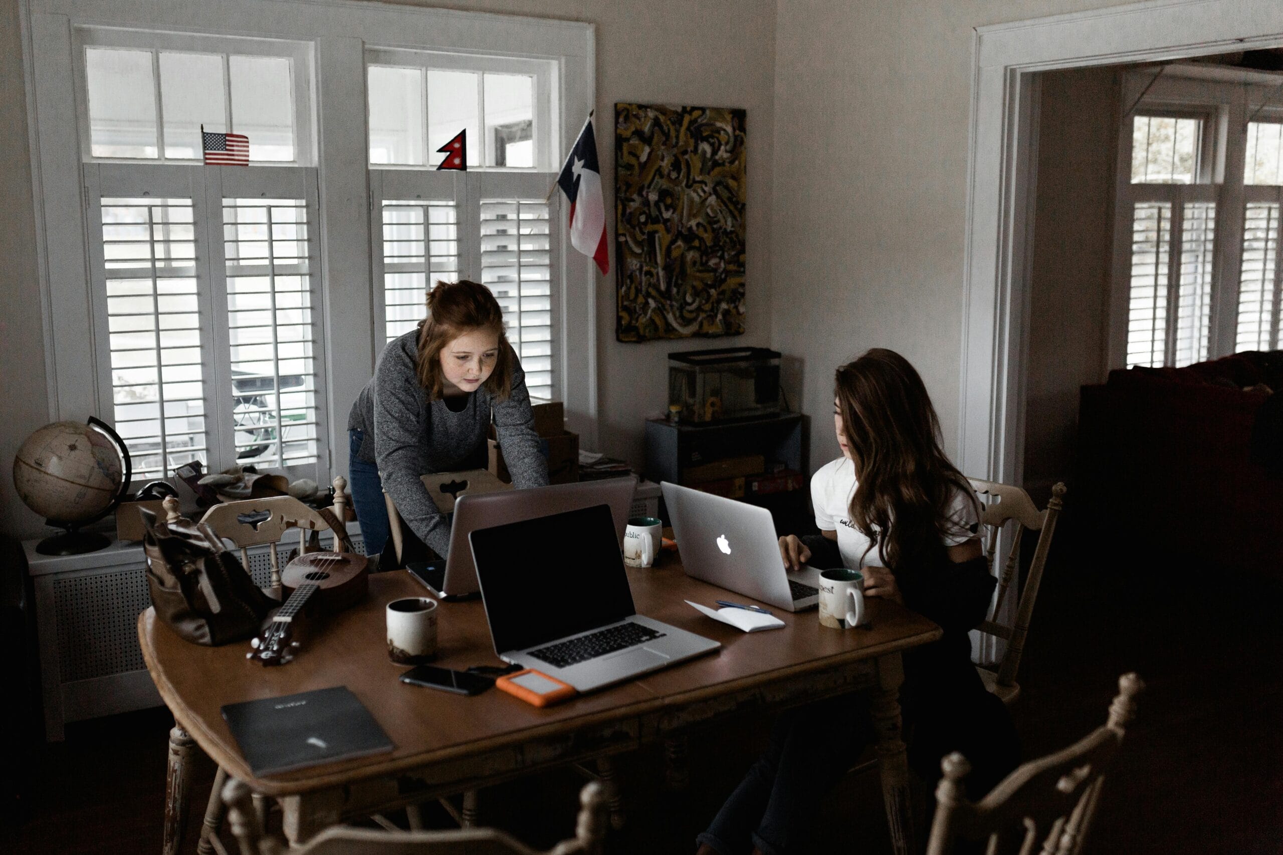 Professional working at a wooden dining table in a cozy room, each using a laptop. The table is scattered with items like a notebook, a globe, a ukulele, mugs, and electronic devices. In the background, flags and an abstract painting add character to the space, with natural light streaming through white shutters on the windows.