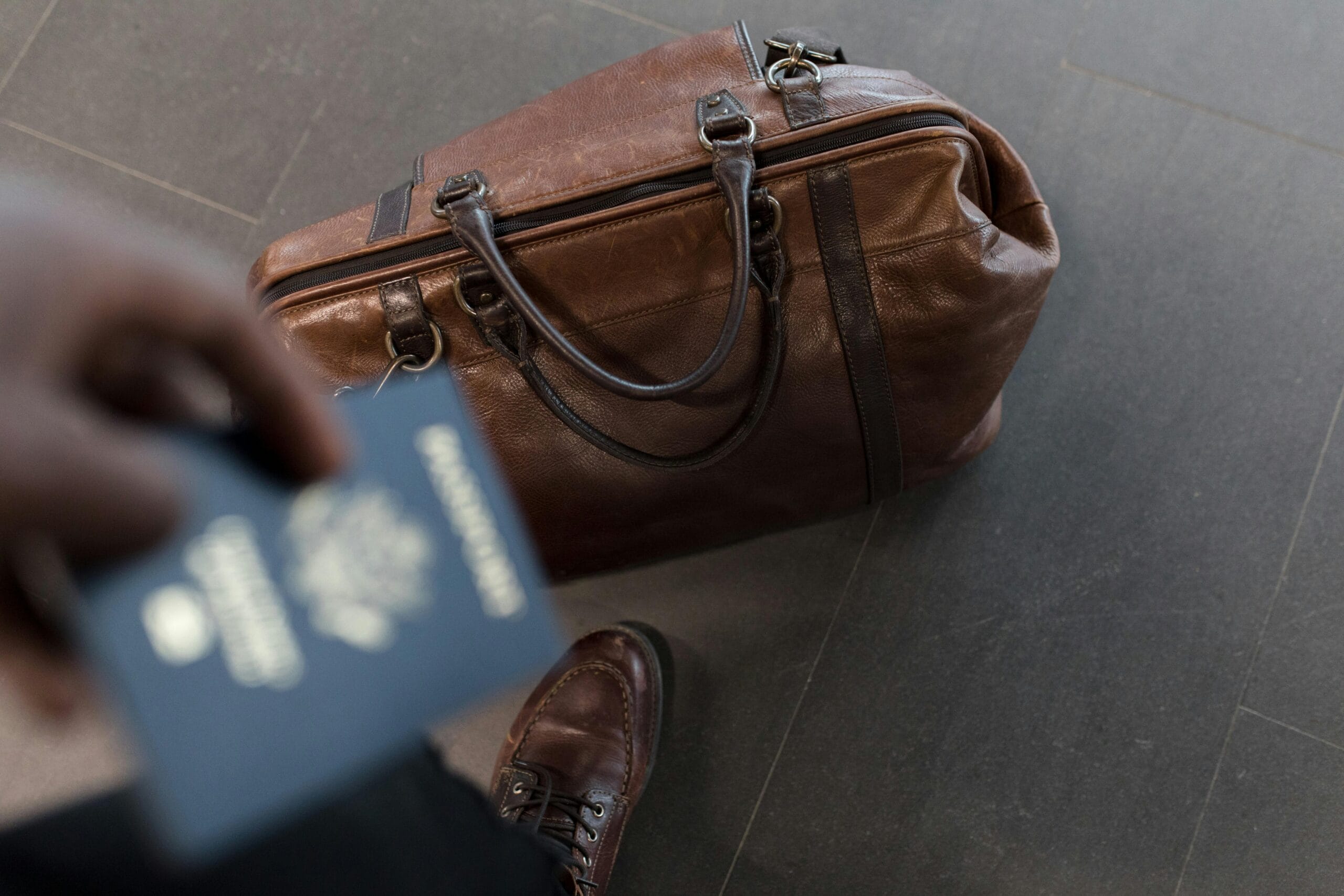 A close-up of a brown leather travel bag placed on a tiled floor, with a person holding a passport in the foreground, ready for their journey