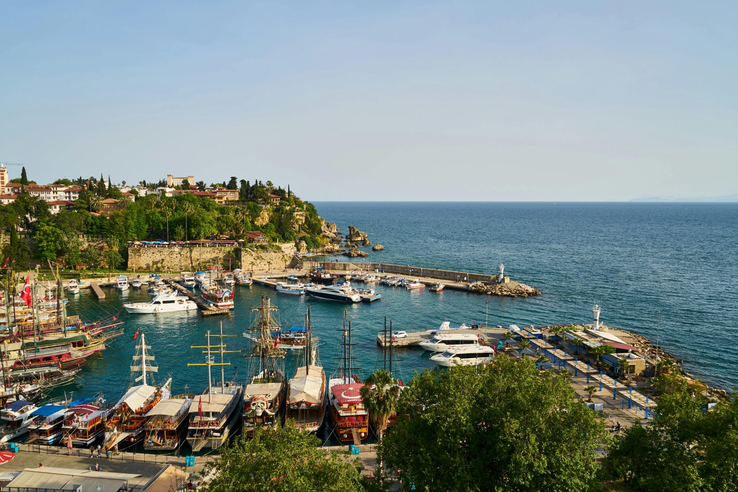 Scenic view of Antalya's Old Town harbor with traditional boats, lush greenery, and the turquoise waters of the Mediterranean Sea under a clear sky.