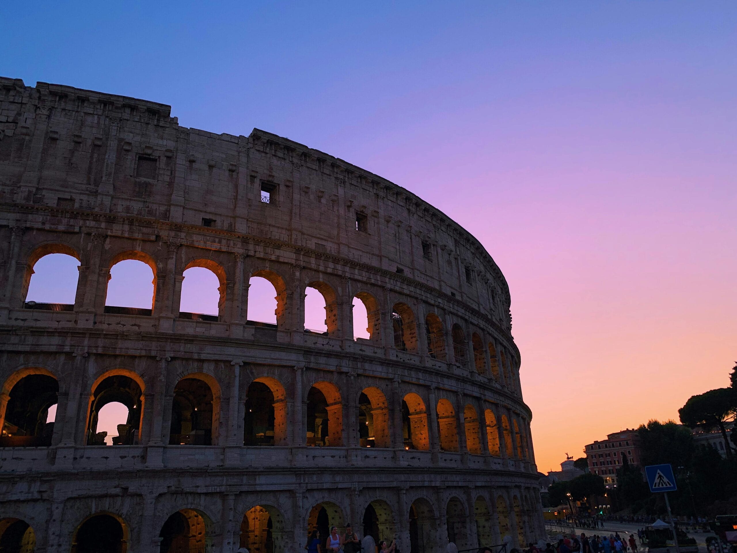 A stunning view of the Colosseum in Rome at sunset, with a vibrant gradient sky transitioning from purple to orange. The iconic arches of the ancient structure are illuminated, creating a beautiful contrast against the darkening surroundings. UK and Europe DMC