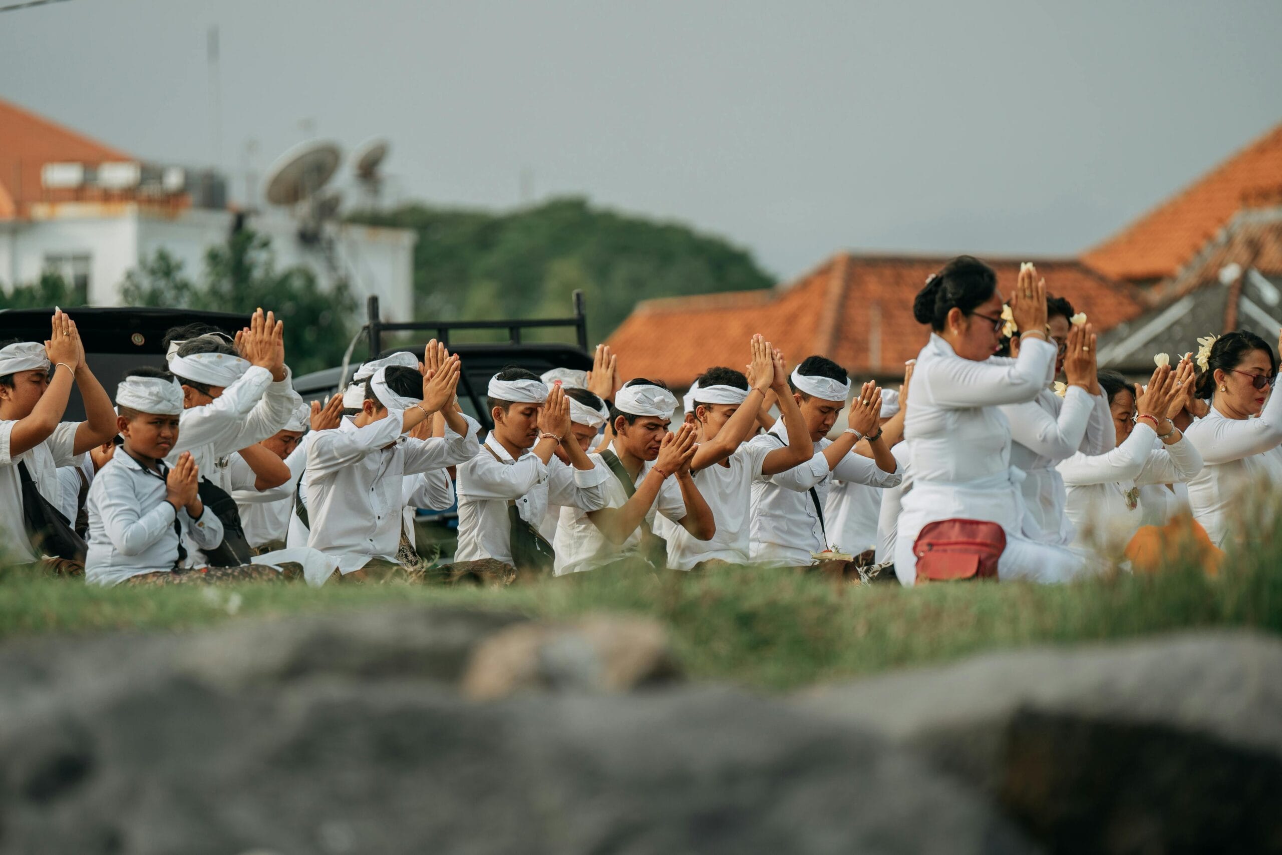 Indonesian people dressed in traditional white attire and headbands participating in a cultural or religious ceremony, seated outdoors with hands clasped in prayer, against the backdrop of tiled rooftops, greenery and and praying in the Street during Nyepi Holiday 