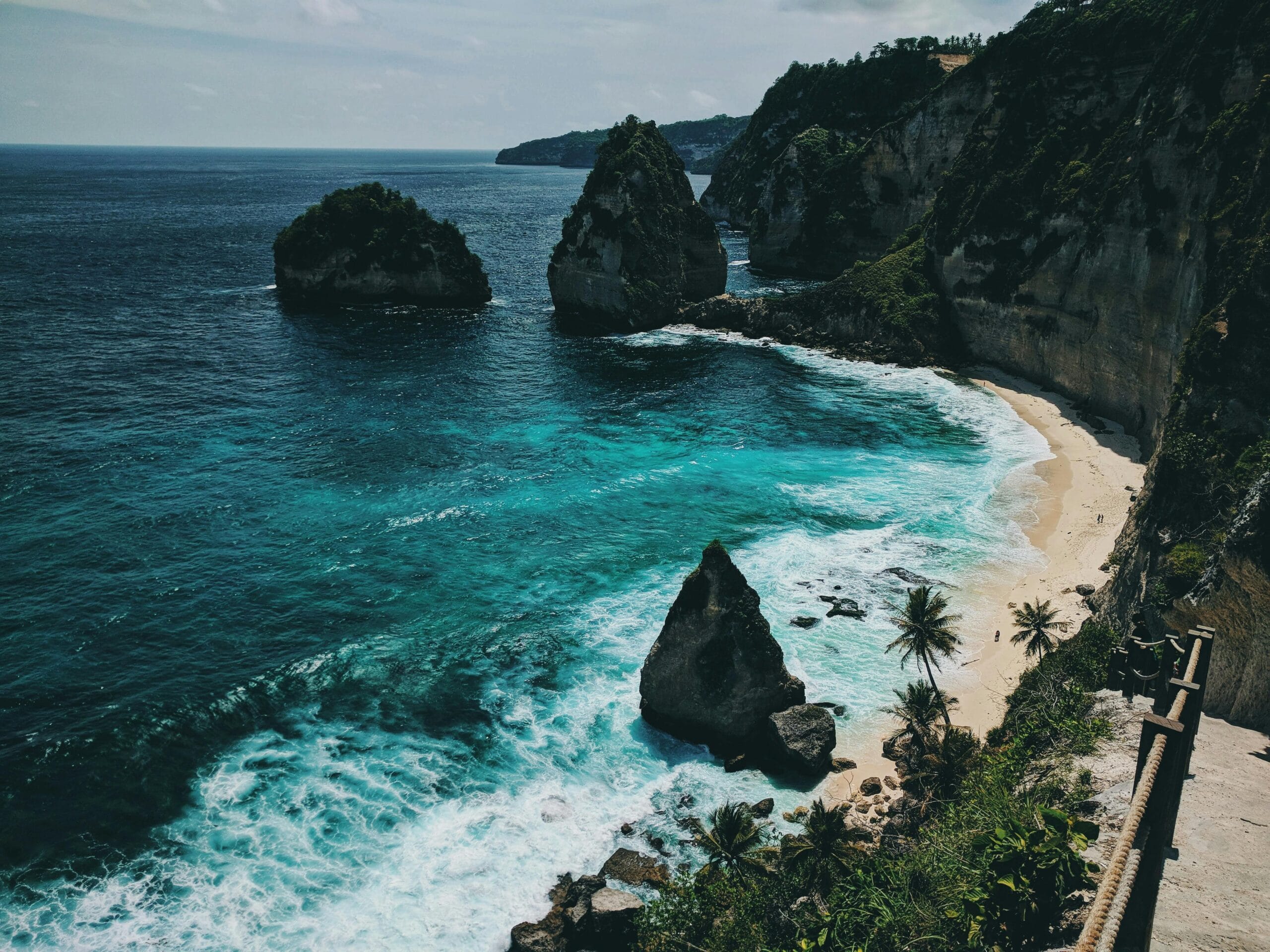 Scenic view of a pristine beach surrounded by towering cliffs and turquoise ocean waves, with lush green vegetation and palm trees lining the landscape. Bali DMC