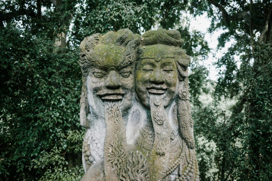 Ancient moss-covered stone sculpture depicting two intricately carved faces with detailed expressions, surrounded by lush green forest foliage. Stone Buddhist Figure in Ubud Monkey Forest , BALI DMC