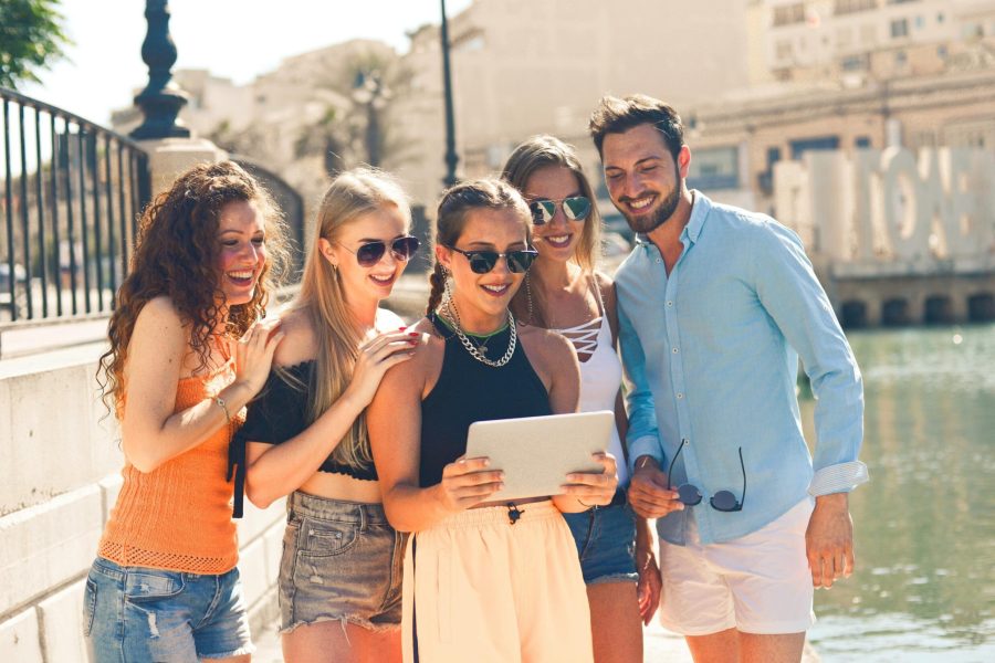 A group of five friends, three women and two men, enjoying a sunny day outdoors near a waterfront. They are gathered around a tablet, smiling and looking at the screen, dressed in casual summer attire, with a cityscape in the background. Europe Group Travel. Group Travel Destinations